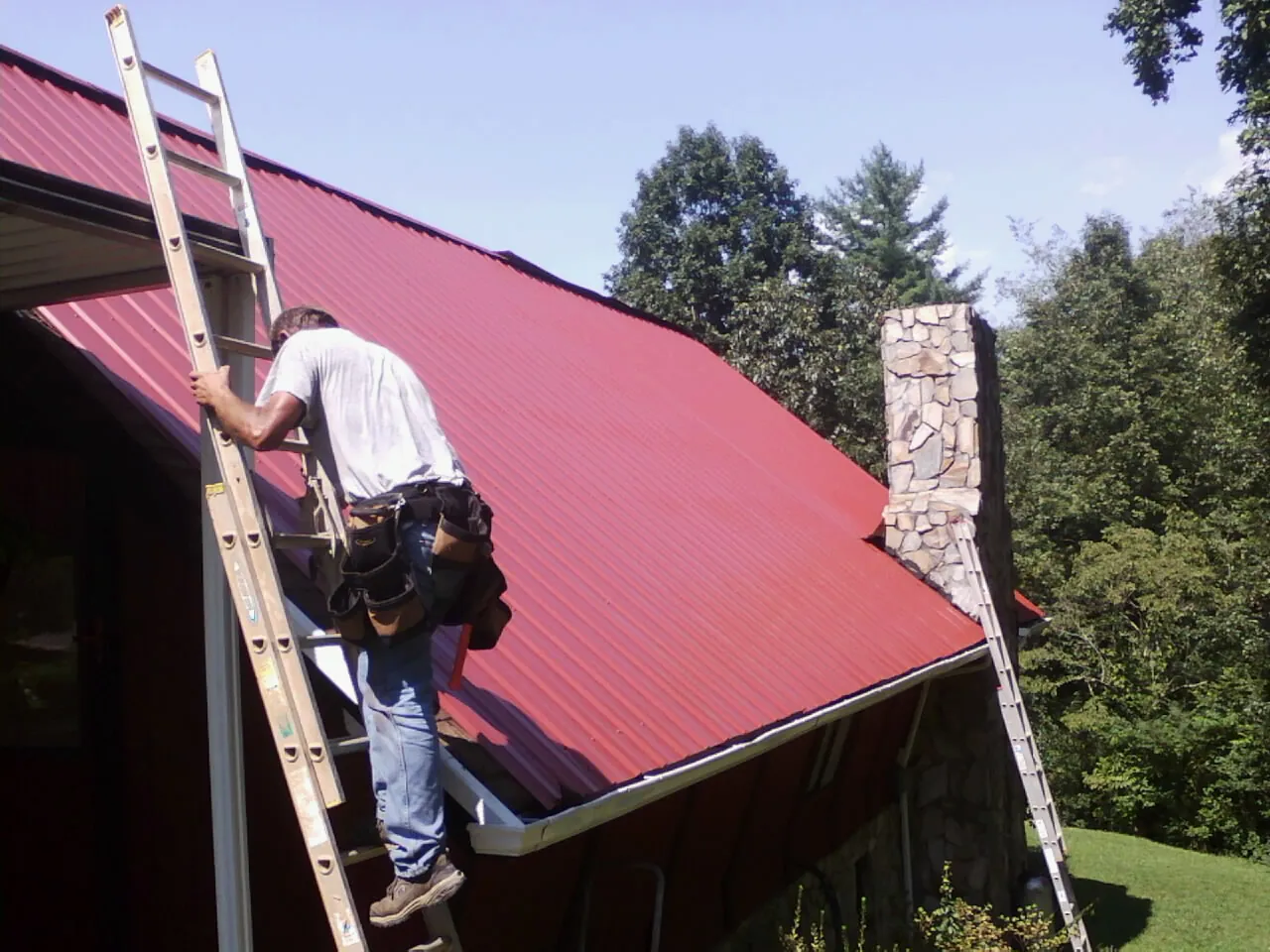 Roofer on a ladder beside a red metal roof and stone chimney — Soffit & Fascia Repair by Roofing Brothers in Supply